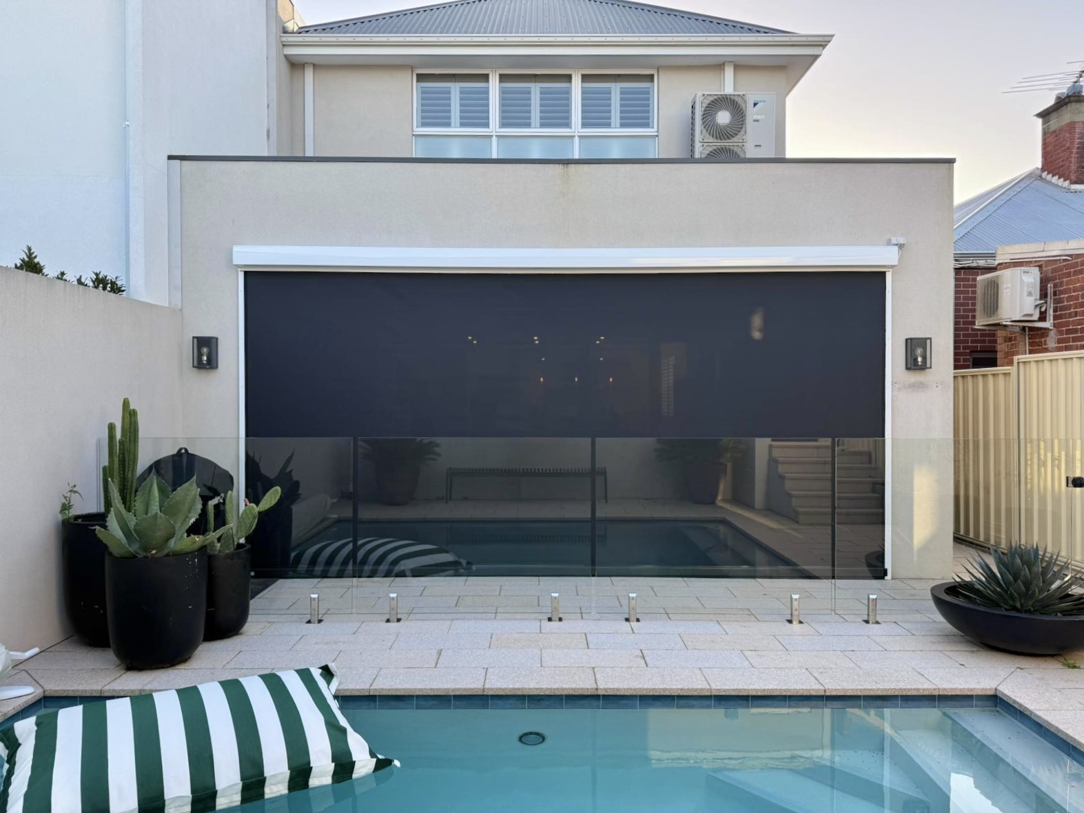 Charcoal Ziptrak blind fully extended over an alfresco area behind a glass-fenced swimming pool at a Perth home, installed by Kenlow